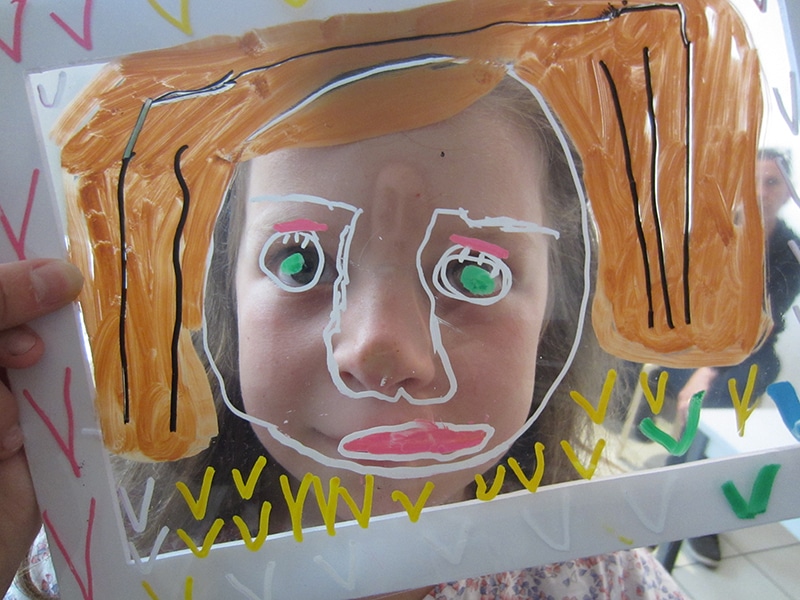 Une enfant a son visage derrière une feuille transparente sur laquelle elle a dessiné un autre visage avec des crayon posca.