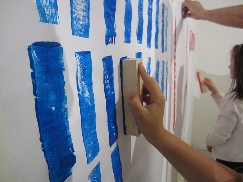 une main d'enfant tient un morceau de bois de forme rectangulaire et s'en sert de tampon pour faire les empreintes sur une feuille blanche au mur. Les empreintes sont réalisées à la peinture bleue et rouge.